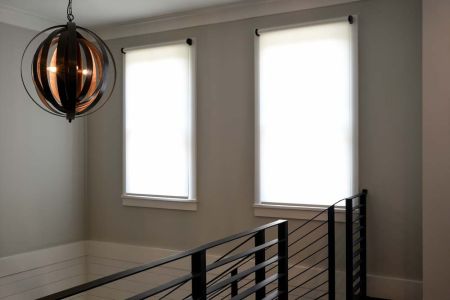 A modern interior hallway featuring two tall windows with white roller shades, a black metal stair railing, and a spherical pendant light fixture with exposed bulbs.