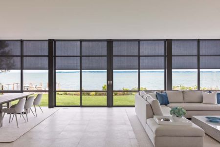 A spacious modern living and dining area with floor-to-ceiling windows overlooking a waterfront view, featuring motorized roller shades, a light gray sectional sofa, and a dining table with white chairs.