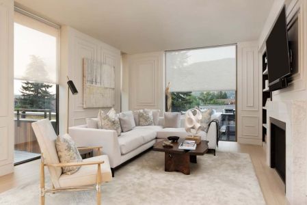 A bright and elegant living room with large windows covered by light-filtering shades, featuring a white sectional sofa, abstract artwork, a wood coffee table, and a wall-mounted TV above a fireplace.