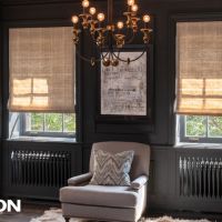 A cozy, elegant living room with dark walls features motorized Lutron shades partially drawn over tall windows, a classic chandelier, and a plush armchair centered below framed artwork.