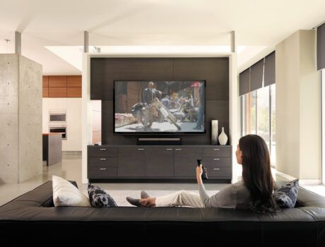 A woman sitting on a couch holding a Savant remote and watching TV. The motorized window shades are partially drawn.