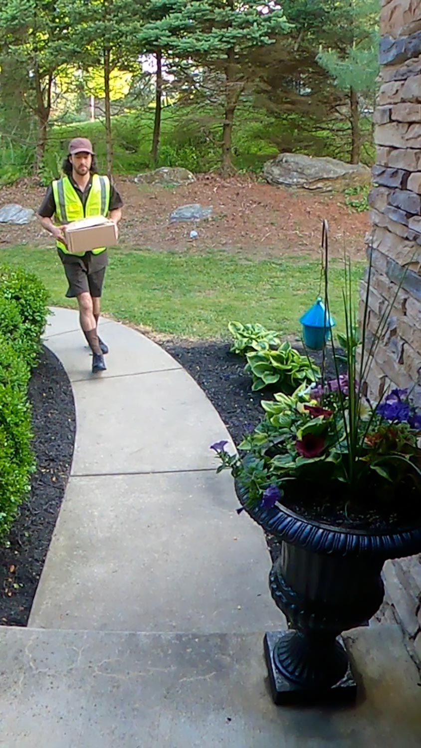 A delivery person in a high-visibility vest walking up a curved front walkway toward the door, holding a package, with landscaped greenery and flowers along the path.