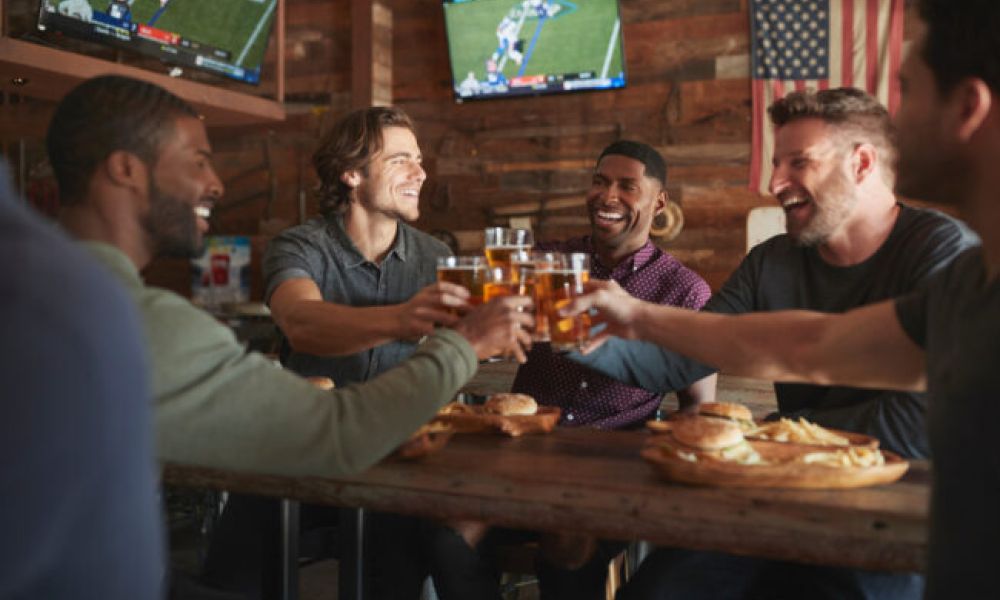 <span class="uk-h4">Group of friends drinking beer at a sports bar with multiple screens.</span>