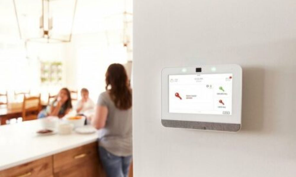 <span class="uk-h4">A kitchen space with a family in the background and an Alarm.com wall panel and interface on the hallway’s wall in the foreground.</span>