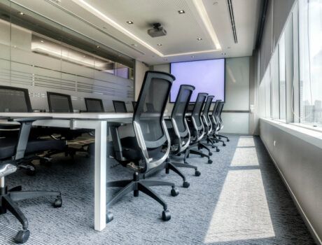 Corporate boardroom with a long conference table, ergonomic chairs, large windows, and a projector screen