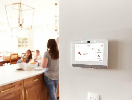 A kitchen space with a family in the background and an Alarm.com wall panel and interface on the hallway’s wall in the foreground.