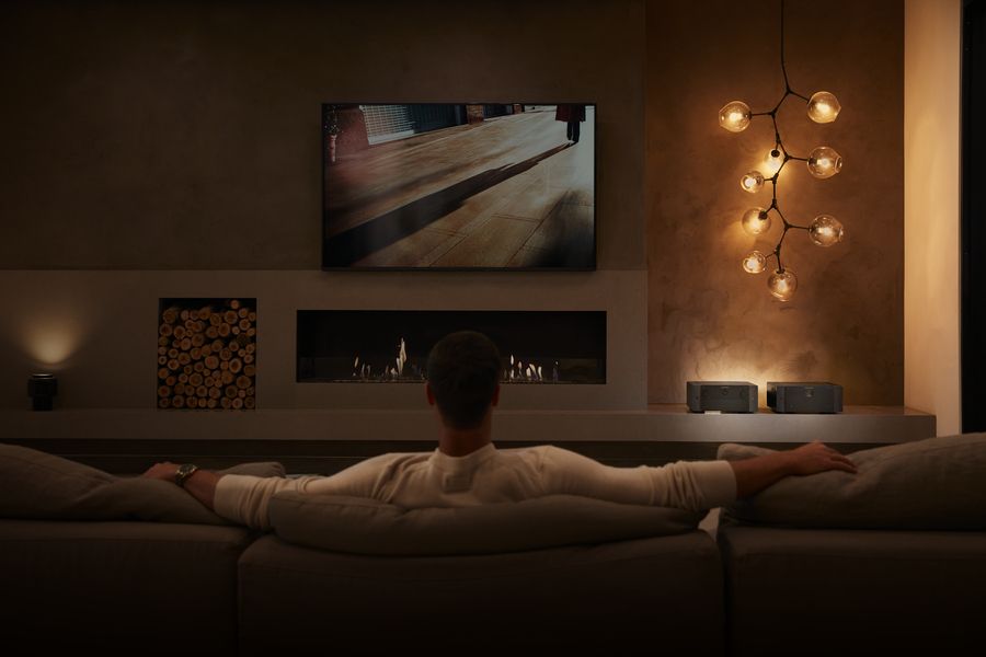 Person sits on couch looking at a large TV display on the wall. The room is dimly lit for enhanced viewing.