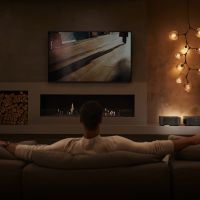 Person sits on couch looking at a large TV display on the wall. The room is dimly lit for enhanced viewing.