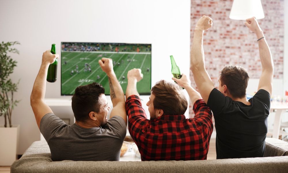 <span class="uk-h4">Three people sitting on a couch watching a football game and cheering on their team.</span>